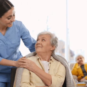 Health and Social Care Management: Caregiver in blue scrubs assisting an elderly person covered with a blanket in a well-lit room, emphasizing professional and compassionate care services