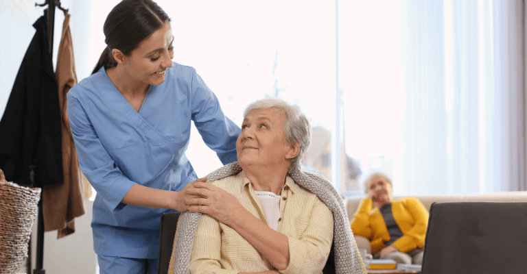 Health and Social Care Management: Caregiver in blue scrubs assisting an elderly person covered with a blanket in a well-lit room, emphasizing professional and compassionate care services