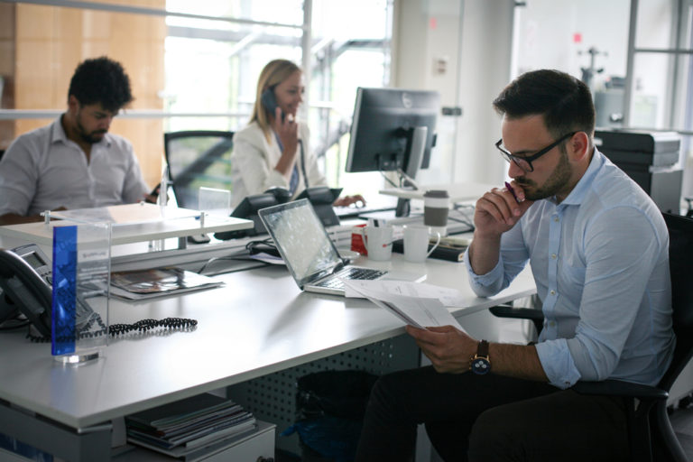 This image captures a modern office setting where three individuals are engaged in various tasks, showcasing practical skills such as multitasking and effective communication. One person is reading a document, another is on the phone, and a third is working on a computer, emphasizing productivity and professional proficiency.