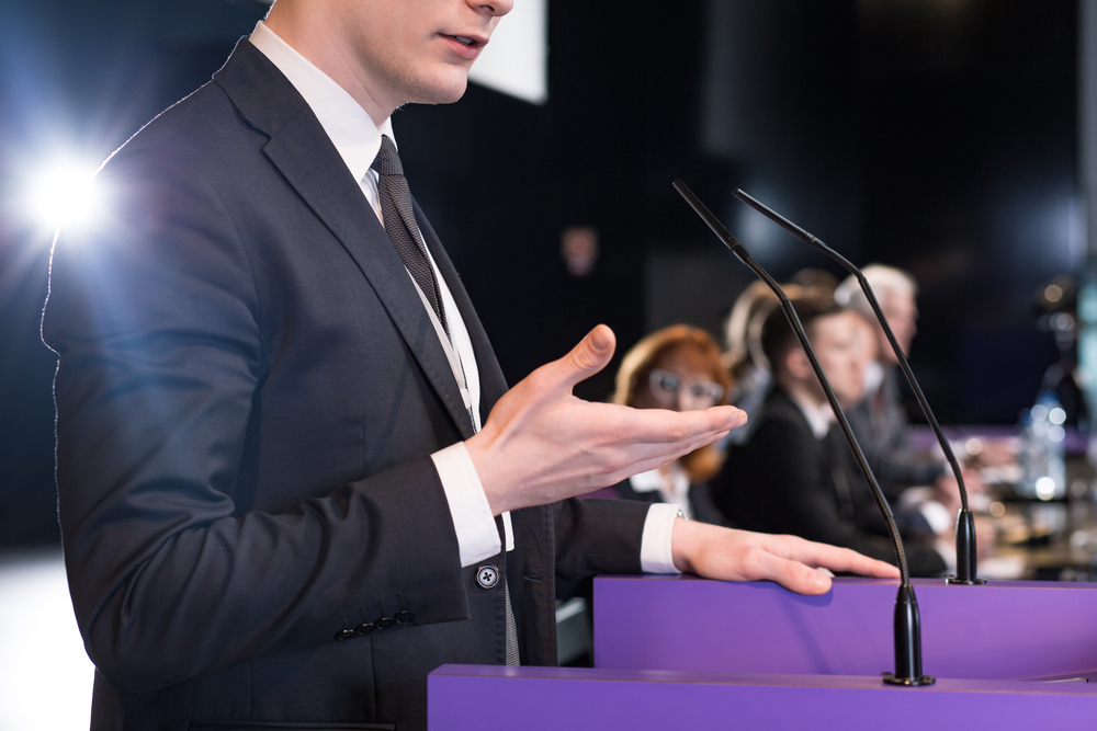 A person in a suit speaks confidently at a podium, gesturing with one hand while addressing an audience. This scene highlights the individual's strong people skills, particularly effective communication and presentation abilities in a formal setting.