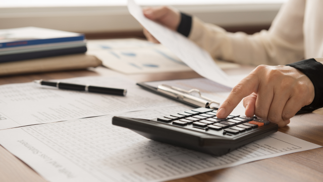 Financial investigator reviewing financial records with a magnifying glass, laptop displaying market data, documents, and cash on a desk, illustrating financial investigation and fraud analysis.