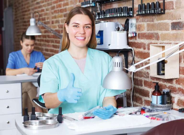 Young happy woman nail master in gloves sitting at her workplace in nail salon