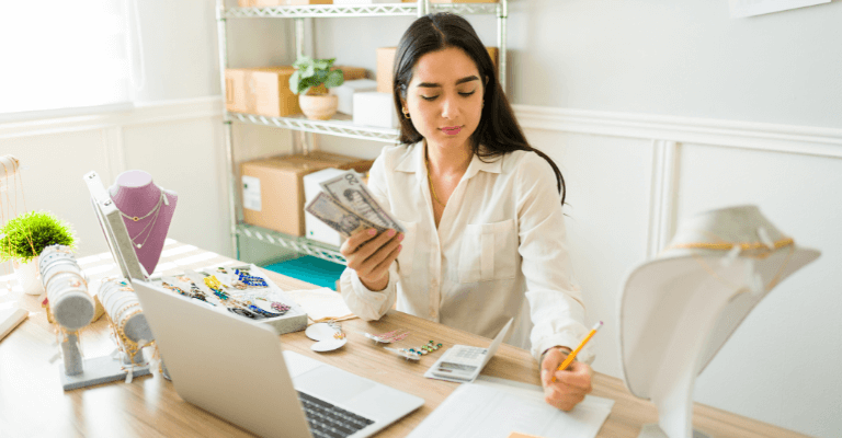Person earning money from home by selling jewelry online, counting cash while working at a desk with a laptop, jewelry displays, and packaging materials.