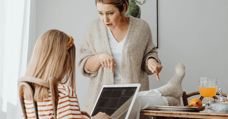 A mother is rudely taunting her daughter, who is sitting with her feet on the dining table. The image represents a discussion on ABC Charts for behavior and their use.