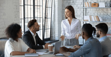 Diverse team in a business meeting with a woman leading the discussion, modern office setting with large windows and bookshelves.