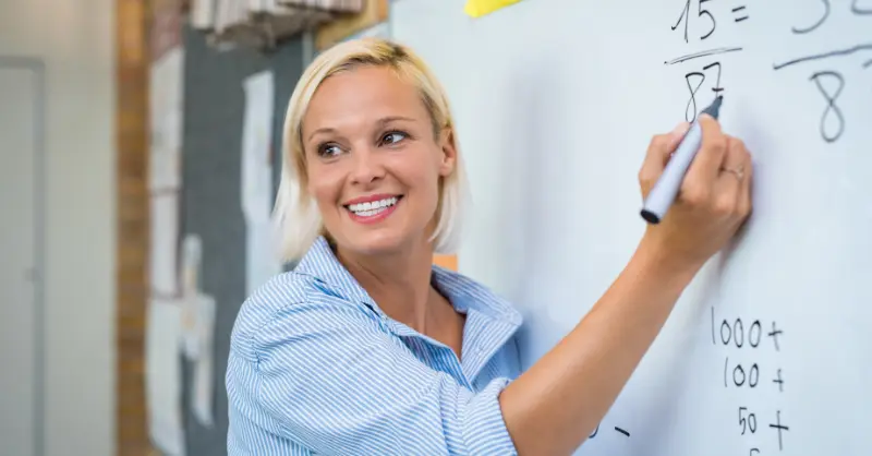 Primary Teacher writing math problems on a whiteboard in a classroom