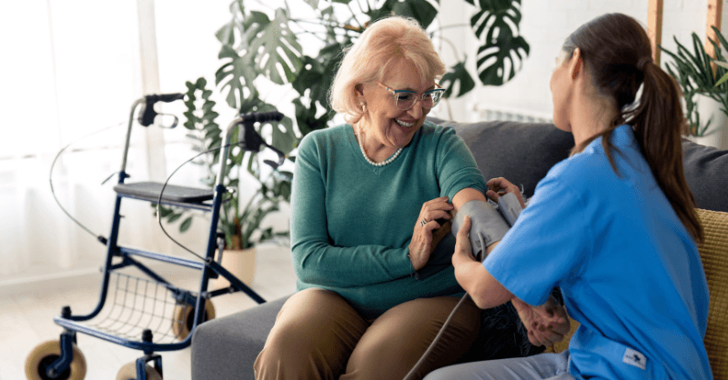 Image showing a healthcare professional in blue scrubs taking the blood pressure of an elderly person sitting on a couch. The elderly person is wearing a green sweater and brown pants. A walker is visible in the background, indicating mobility assistance. The setting appears to be a home environment with plants and natural light. Why Primary Care Matters This image highlights the importance of primary care in providing in-home medical support for elderly individuals, ensuring their well-being and addressing their health needs.