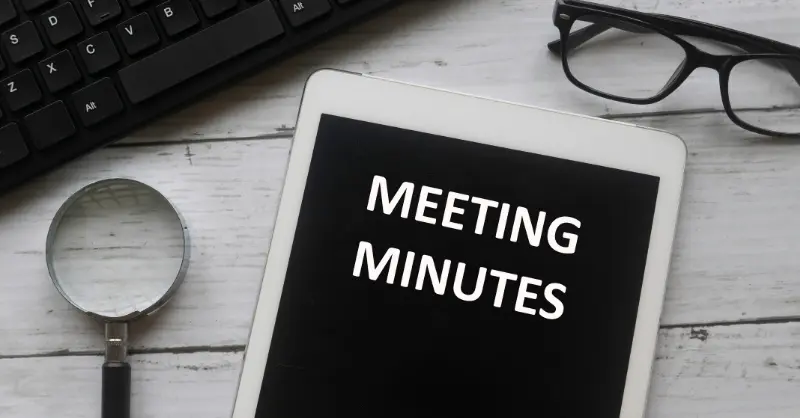 Minute taking setup showing tablet with 'Meeting Minutes' on screen, magnifying glass, eyeglasses, and keyboard on a desk
