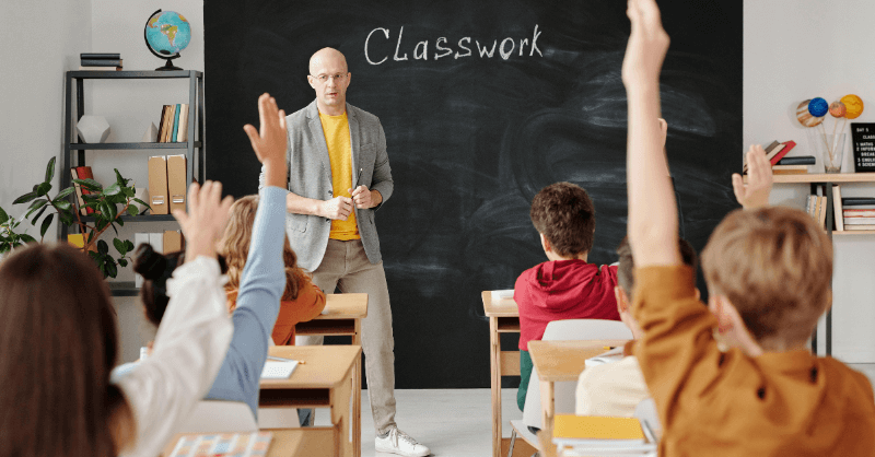 Engaged classroom with students raising hands and a teacher leading 'Classwork' is ideal for illustrating how to get started in teaching without a degree